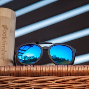 May include: Black sunglasses with blue mirrored lenses, resting on a wicker basket. The sunglasses have a thin metal frame and the brand name "oaktimber" is visible on a bamboo case.