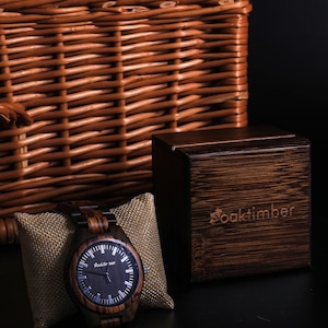 May include: A dark brown wooden wristwatch with a black face and silver hands. The watch is displayed on a small brown pillow and a wooden box with the word "Oaktimber" printed on it.