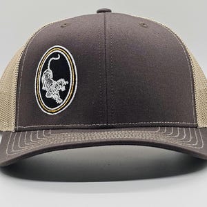 May include: A dark gray trucker hat with a tan mesh back. The front of the hat features an embroidered tiger design within an oval frame. The brim is dark gray with white stitching.