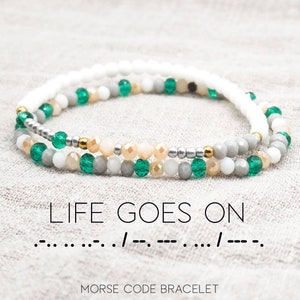 May include: A white, gray, and green beaded bracelet with the message "LIFE GOES ON" in Morse code. The bracelet is made of small, round beads and has a delicate, feminine look.