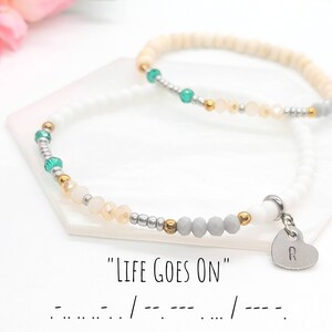 May include: Two beaded bracelets. One is white with gray, gold, and green beads, and a heart charm with the letter "R". The other is beige with green and gold beads. The text "LIFE GOES ON" is below the bracelets.