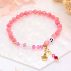 May include: Pink gemstone bracelet with a gold paw print charm and a letter "D" charm. The bracelet is made with small, faceted pink beads and a few clear beads.