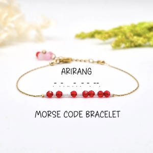 May include: A delicate gold-toned bracelet with red and white beads. The bracelet spells out "Arirang" in Morse code. The words "Morse Code Bracelet" are written below the bracelet in black script.