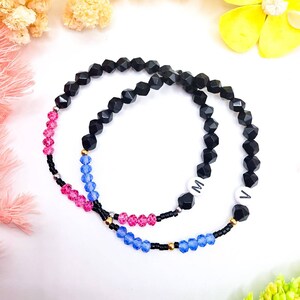 May include: Two beaded bracelets. One bracelet features black, pink, and blue beads, with the letters "M" and "V" on white beads. The other bracelet is primarily black with pink and blue accents. Both bracelets have a delicate, handcrafted appearance.