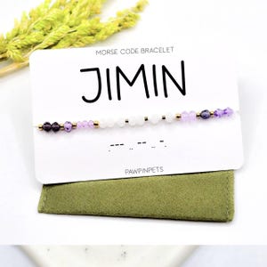 May include: A Morse code bracelet featuring purple, lavender, and white beads with small gold-tone spacers on a white display card. The card displays the text JIMIN, the words MORSE CODE BRACELET, the Morse code sequence, and the brand name PAWPINPETS.