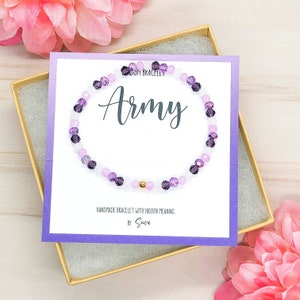 May include: A purple and white beaded bracelet with a gold bead in the center. The bracelet is displayed in a purple box with a white card that says "Army" and "Handmade bracelet with hidden meaning. By Swoon."