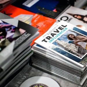 Puede incluir: Una pila de revistas con una revista en la parte superior titulada "Travel Influencer: Gu&iacute;a paso a paso para convertirte en un influencer de viajes". La portada de la revista presenta a una mujer con una camisa blanca y un sombrero de paja, sosteniendo un tel&eacute;fono y sonriendo.