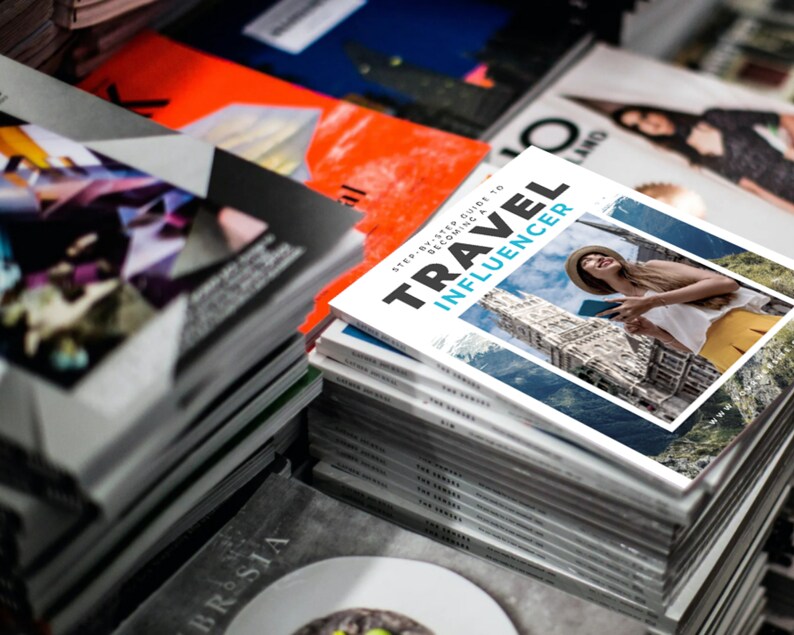 Puede incluir: Una pila de revistas con una revista en la parte superior titulada "Travel Influencer: Gu&iacute;a paso a paso para convertirte en un influencer de viajes". La portada de la revista presenta a una mujer con una camisa blanca y un sombrero de paja, sosteniendo un tel&eacute;fono y sonriendo.