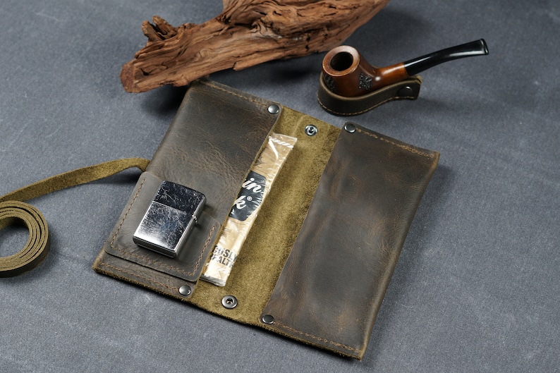 Pu&ograve; includere: Una custodia per tabacco in pelle marrone con fodera verde, un accendino argentato e un pacchetto di tabacco. La custodia ha una cinghia e si chiude con bottoni a pressione.