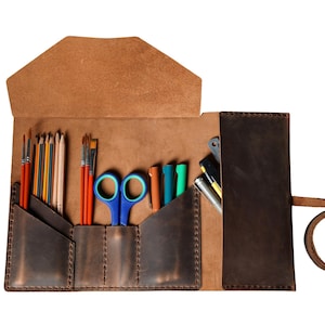 May include: Open leather art supply roll with various tools. Includes paint brushes, pencils, scissors, and pens. The roll is brown with a flap closure and a strap for securing. The interior pockets are visible.