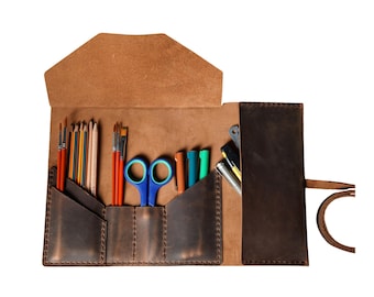 May include: Open leather art supply roll with various tools. Includes paint brushes, pencils, scissors, and pens. The roll is brown with a flap closure and a strap for securing. The interior pockets are visible.