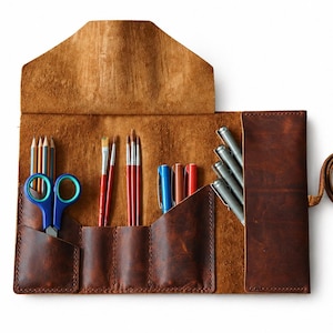 May include: A brown leather artist's roll-up case, open to display art supplies. The case holds pencils, blue scissors, paint brushes, pens, and colored pencils. A leather strap is attached for securing the case.