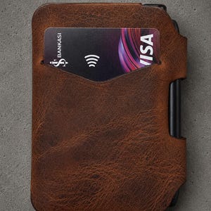 May include: A brown textured leather card holder with a black metal frame edge. A black payment card is inserted into the front slot, displaying the text Bankasi and Visa along with a contactless payment symbol on the right side of the card.