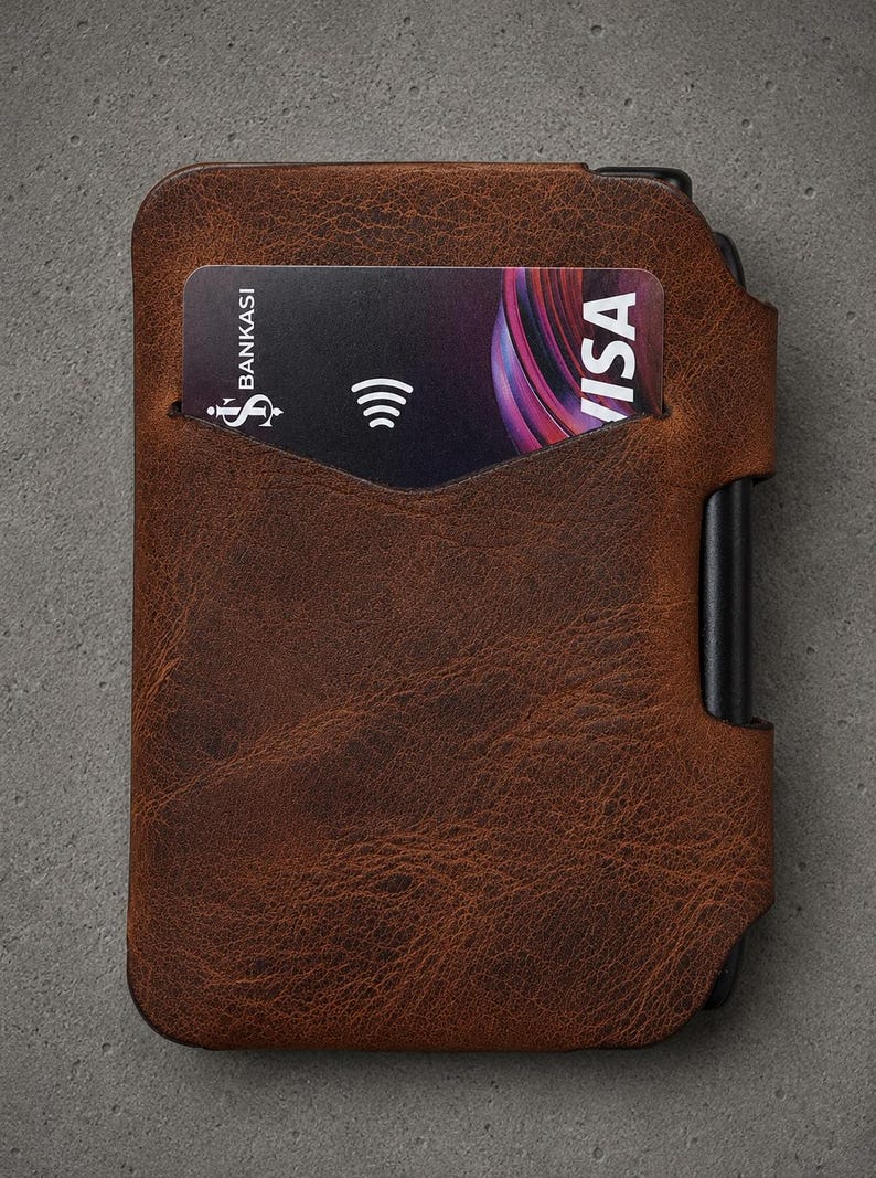 May include: A brown textured leather card holder with a black metal frame edge. A black payment card is inserted into the front slot, displaying the text Bankasi and Visa along with a contactless payment symbol on the right side of the card.