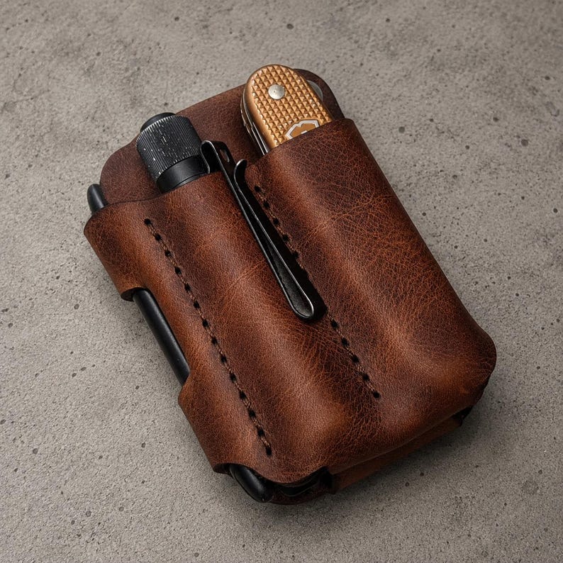 May include: A brown leather pocket organizer featuring two vertical slots. One slot holds a black metal pen with a pocket clip, while the other slot contains a small folding pocket knife with a textured gold-colored handle and a silver rivet.