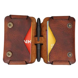 Op de afbeelding: Open bruin leren portemonnee met twee kaartsleuven, elk met een creditcard. Een zwarte pen staat in het midden. De portemonnee heeft aan elke kant een drukknoopsluiting. Het leer heeft een distressed look.