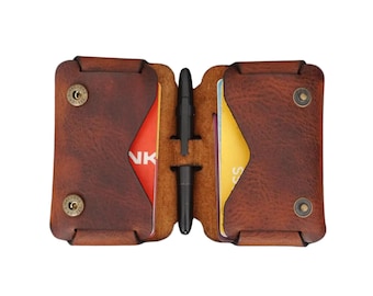 May include: Open brown leather wallet with two card slots, each holding a credit card. A black pen is in the centre. The wallet has a snap closure on each side. The leather has a distressed look.