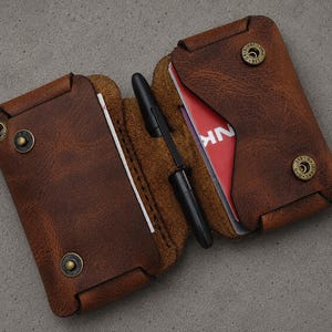 May include: A brown leather wallet with a textured finish, featuring two main compartments secured by brass-colored metal snaps. A black pen is held in the center fold. The wallet contains visible white and red cards with partial text.
