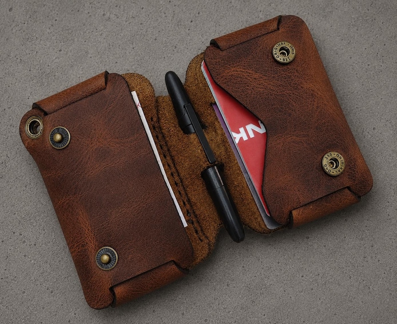 May include: A brown leather wallet with a textured finish, featuring two main compartments secured by brass-colored metal snaps. A black pen is held in the center fold. The wallet contains visible white and red cards with partial text.