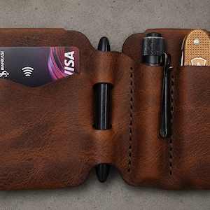 May include: A brown leather pocket organizer featuring a dedicated slot for a credit card, a central loop holding a black pen, and two additional vertical sleeves containing a small black flashlight and a gold-colored folding pocket knife.