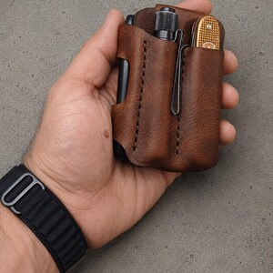 May include: A brown leather pocket organizer featuring two vertical slots. One slot holds a small black flashlight with a metal clip, and the other slot contains a gold-colored folding pocket knife with a textured handle and a metal clip.