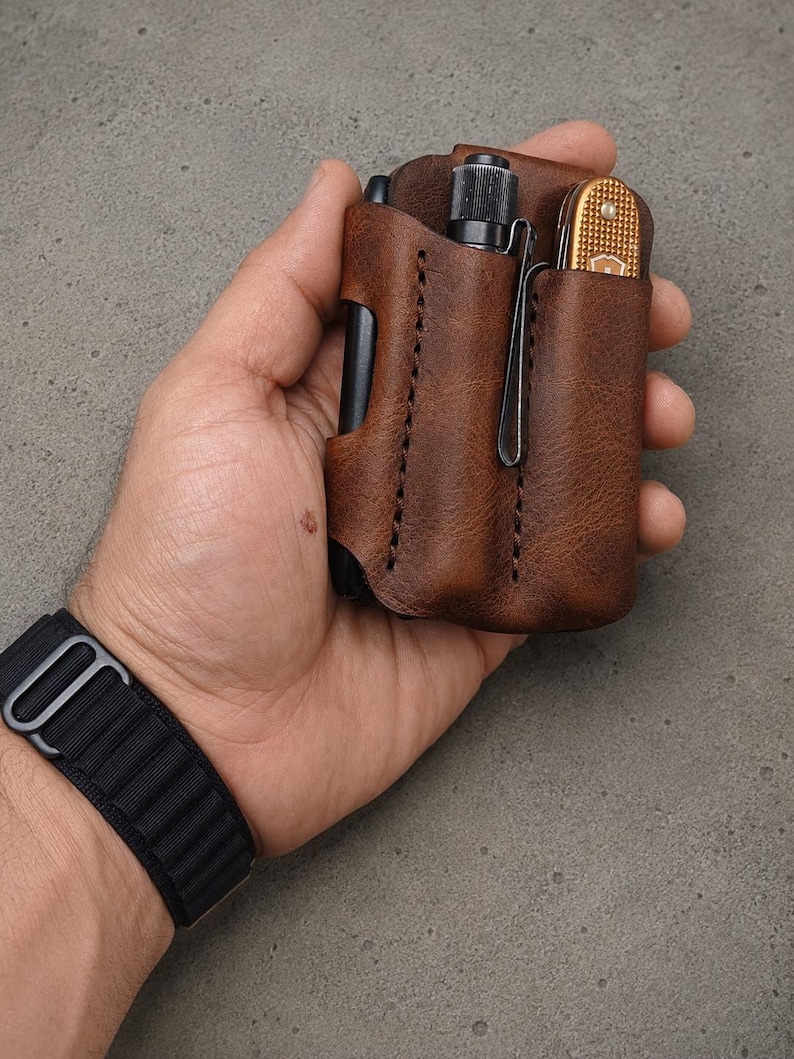 May include: A brown leather pocket organizer featuring two vertical slots. One slot holds a small black flashlight with a metal clip, and the other slot contains a gold-colored folding pocket knife with a textured handle and a metal clip.