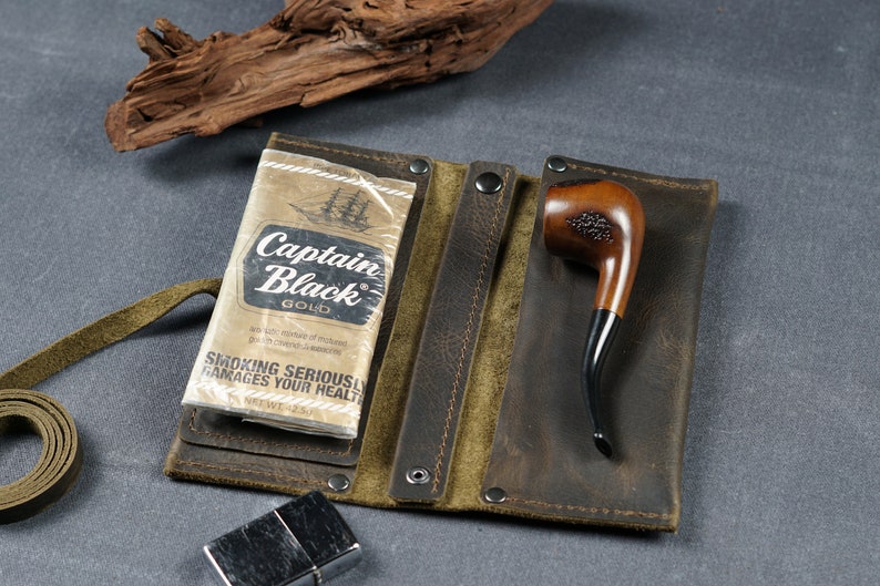 Pu&ograve; includere: Una custodia in pelle marrone per tabacco con chiusura a zip. La custodia ha due scomparti, uno per un pacchetto di sigarette Captain Black Gold e l'altro per una pipa di legno. La custodia ha anche un cinturino in pelle verde.