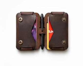 May include: Open brown leather wallet with two sections, each secured by a snap closure. Inside, there are credit cards and a black pen. The wallet is on a white background.