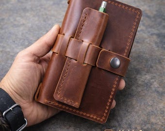 May include: A brown leather notebook cover with a pen holder and press stud closure. The cover has a stitched design and holds three notebooks. The pen is green and silver. The leather is a warm, medium brown colour.