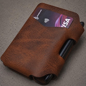 May include: A brown textured leather wallet featuring a card slot containing a Bankasi Visa card with a contactless payment symbol. A black pen is inserted into a dedicated side loop integrated into the structure of the wallet casing.