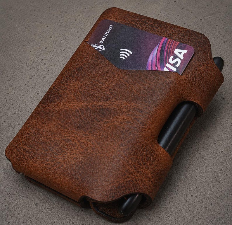 May include: A brown textured leather wallet featuring a card slot containing a Bankasi Visa card with a contactless payment symbol. A black pen is inserted into a dedicated side loop integrated into the structure of the wallet casing.