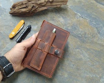 May include: A brown leather notebook with a strap and button closure. The notebook has a pen holder and is made of distressed leather.