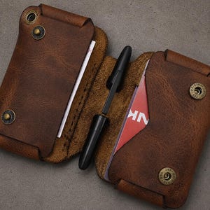 May include: A brown leather wallet with two main compartments secured by brass-colored metal snap buttons. The wallet holds white paper and a red card with visible white text. A black pen is positioned vertically in the center of the wallet.