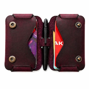 May include: An open burgundy leather wallet with a black pen and cards visible inside. The wallet features brass-coloured snap closures. The interior is a lighter shade of burgundy.