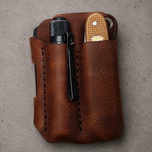 May include: A brown leather pocket organizer featuring two vertical slots. The left slot holds a black cylindrical flashlight with a metal clip, while the right slot contains a tan multi-tool with a textured grip and a visible metal ring attachment.