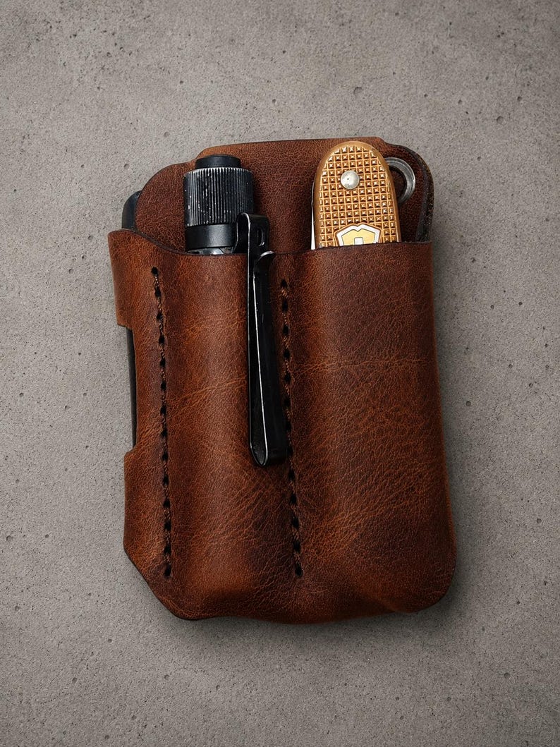 May include: A brown leather pocket organizer featuring two vertical slots. The left slot holds a black cylindrical flashlight with a metal clip, while the right slot contains a tan multi-tool with a textured grip and a visible metal ring attachment.