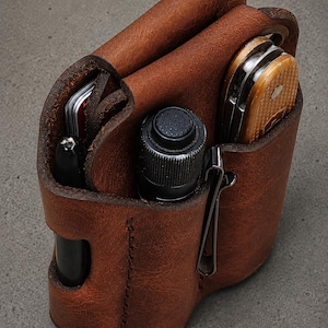 May include: A brown leather multi-tool organizer pouch featuring three distinct vertical compartments. It holds a black pen, a small black cylindrical flashlight with a textured button, and a folding pocket knife with a tan handle.