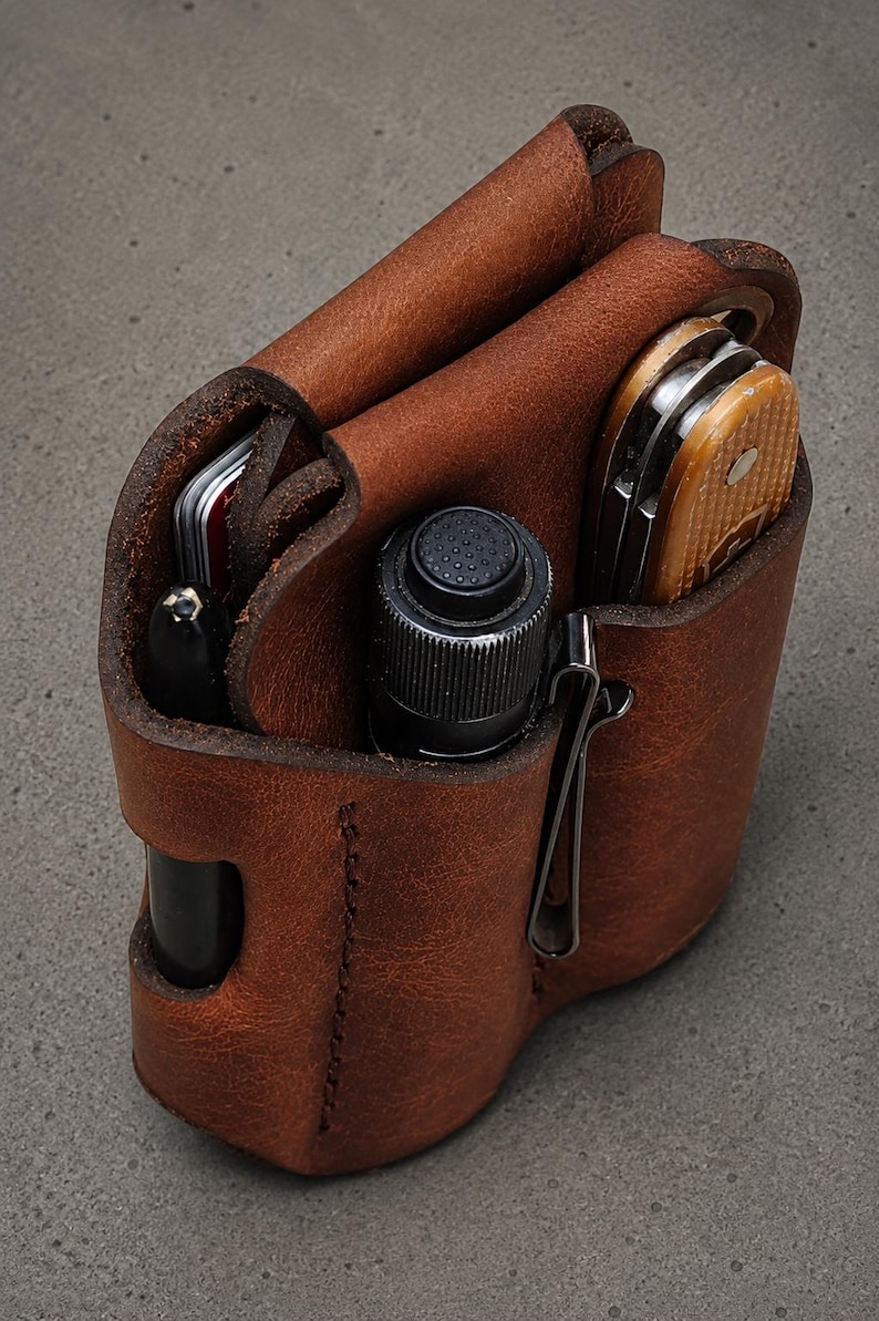 May include: A brown leather multi-tool organizer pouch featuring three distinct vertical compartments. It holds a black pen, a small black cylindrical flashlight with a textured button, and a folding pocket knife with a tan handle.