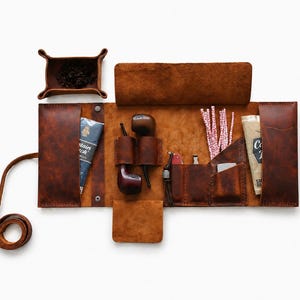 May include: A brown leather tobacco pipe roll, open to display its contents. The roll includes a tobacco tray, two pipes, a pouch, and other accessories. The leather is a rich, warm brown color, and the roll is designed to be portable.