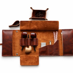 May include: A brown leather tobacco pipe roll with multiple compartments. The roll contains two pipes, a lighter, a small bottle, pipe cleaners, and a tobacco tray. The roll is open, revealing the contents, and is set against a white background.