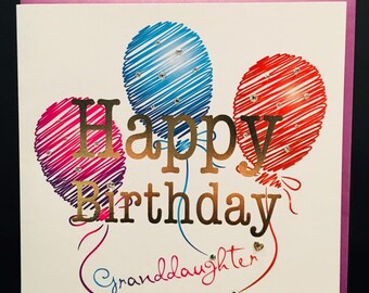 May include: A white card with a pink border. The card has a gold foil inscription that reads "Happy Birthday Granddaughter". The inscription is surrounded by three colorful balloons with glitter accents.
