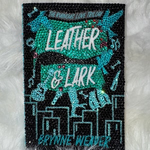 Puede incluir: Una portada del libro "Leather & Lark" de la serie "The Ruinous Love Trilogy", adornada con pedrería negra, turquesa y roja. El título y el nombre del autor son visibles. Se presentan notas musicales y siluetas.