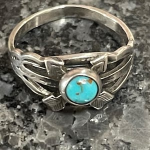 May include: A silver ring with a turquoise stone set in the center. The ring has an openwork design with leaf and arrow-like details. The turquoise stone is round and a vibrant blue-green color. The ring is likely a piece of jewelry.