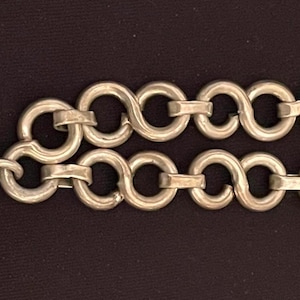 May include: Silver-toned bracelet featuring a decorative, stylized clasp and a double-strand chain. The clasp has a rounded central element and a curved, embellished border. The chain is composed of interlocking loops, and the bracelet has a T-bar closure.