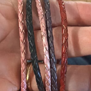 May include: A collection of six braided leather bracelets in various colors, including pink, black, mauve, and red. The bracelets are displayed close-up, showcasing the texture and detail of the woven design. The bracelets are likely for wrist wear.