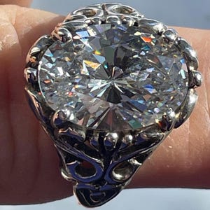 May include: A large, ornate silver ring featuring a massive, clear, faceted gemstone. The ring's setting is detailed with intricate scrollwork and decorative elements, creating a bold and eye-catching design. The gemstone sparkles with internal reflections.