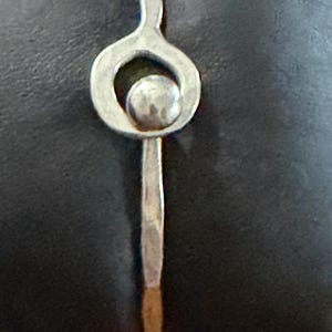 May include: A silver-toned bracelet with a unique design. The bracelet features a circular element with a small sphere in the center, connected to a slender, textured band. The metal has a hammered appearance, giving it a handcrafted look.