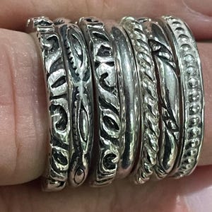 May include: A collection of silver rings with various designs, including textured patterns, rope-like twists, and beaded edges. The rings are stacked together, showcasing the different styles and textures of each piece. The rings are worn on a finger.