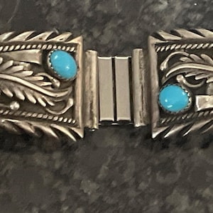 May include: Silver-toned bracelet with intricate leaf and scroll designs. Each section features a vibrant turquoise and a red stone. The bracelet is connected by a silver clasp, showcasing a Southwestern style.