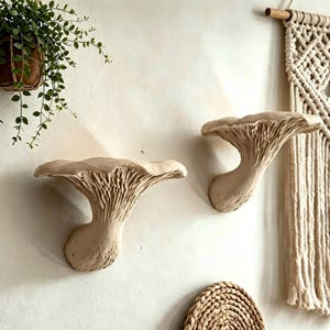 Può includere: Due mensole da parete color beige a forma di fungo con sottostanti testurizzati simili a lamelle. Queste unità scultoree montate a parete presentano una superficie superiore ampia e piatta e una base curva e affusolata.